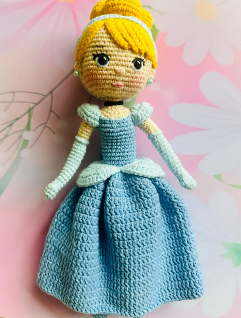 May include: A handmade crochet doll depicting a princess in a blue dress. The doll has yellow hair styled in a bun, blue eyes, and a light skin tone. The dress features a detailed crochet pattern and light blue accents.