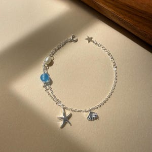 Puede incluir: Una pulsera de cadena plateada que presenta una pequeña perla blanca, una cuenta azul claro translúcida, un dije de estrella de mar plateado, un dije de concha marina plateado y una pequeña estrella plateada en el cierre.