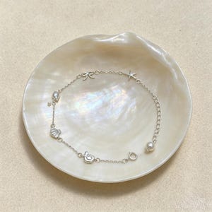 May include: A silver-toned charm bracelet featuring a delicate chain with small charms including a bow, a starfish, a seashell, and a spiral shell, accented with two small white pearls and a circular clasp closure with an adjustable chain link.