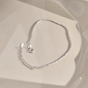 May include: A silver-toned bracelet featuring a textured, sparkling chain design. The piece includes a circular spring ring clasp and an adjustable extender chain with a small, flat, oval-shaped tag stamped with the text S925 for authenticity.