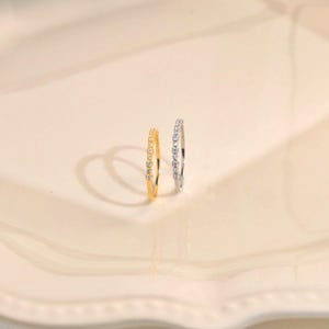 Könnte beinhalten: Zwei dünne Ringe, einer in Goldoptik und einer in Silberoptik, jeweils mit einer Reihe kleiner, klarer, rund geschliffener Steine entlang der Ringschiene. Die Ringe stehen aufrecht und zeigen ihre Kreisform sowie die funkelnden Steindetails.