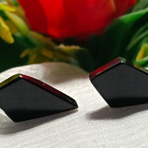 May include: A pair of black stud earrings with a geometric, asymmetrical, multi-faceted shape. The earrings have a smooth, polished surface and a sharp, angular silhouette, presenting a dark, solid colour throughout each individual piece.