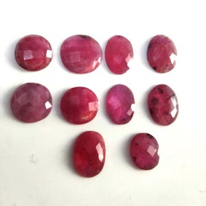 May include: A collection of ten faceted, oval and round loose gemstones in varying shades of deep pink and magenta. Each stone features a polished, multi-faceted surface that reflects light, displaying natural inclusions and unique color variations.