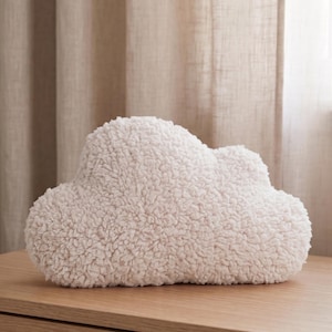 May include: A soft, off-white cloud-shaped cushion with a textured, fluffy surface. The cushion sits on a light-coloured wooden surface. The background features neutral-toned curtains, creating a calming aesthetic.