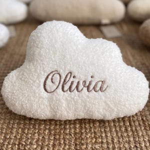 May include: A white cloud-shaped cushion embroidered with the name "Olivia" in brown thread. The cushion has a soft, textured surface, with other cloud cushions in the background. The surface is a woven, brown material.