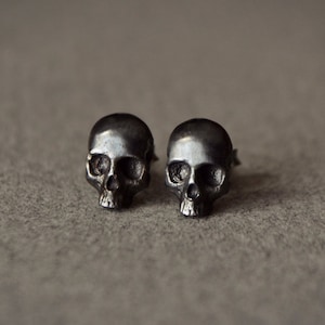 Può includere: Un paio di orecchini a bottone a forma di teschio in argento. I teschi sono dettagliati e hanno una finitura leggermente testurizzata.