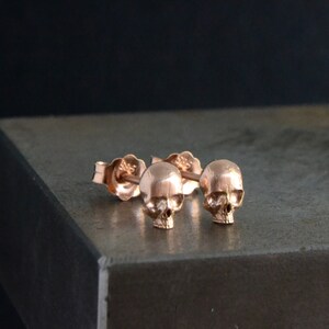 May include: A pair of rose gold skull stud earrings. The earrings are small and delicate, and they feature a detailed skull design.