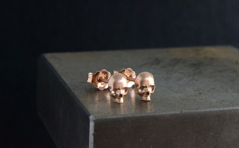 May include: A pair of rose gold skull stud earrings. The earrings are small and delicate, and they feature a detailed skull design.