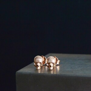 May include: A pair of rose gold skull stud earrings. The earrings are small and delicate, and they have a simple, minimalist design.