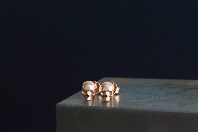May include: A pair of rose gold skull stud earrings. The earrings are small and delicate, and they have a simple, minimalist design.