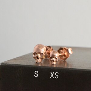 May include: Two rose gold skull stud earrings, one larger than the other. The larger earring is labeled "S" and the smaller earring is labeled "XS".