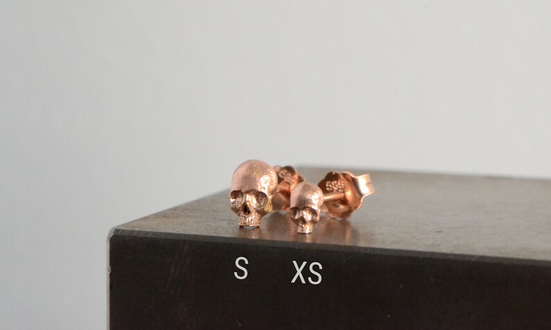 May include: Two rose gold skull stud earrings, one larger than the other. The larger earring is labeled "S" and the smaller earring is labeled "XS".