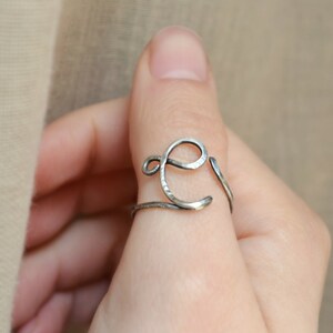 May include: A silver ring with a hammered finish, shaped like the letter E, worn on a finger.
