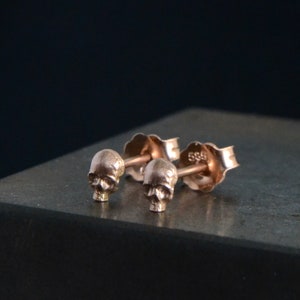 May include: A pair of rose gold skull stud earrings. The earrings are small and delicate, and they feature a detailed skull design.