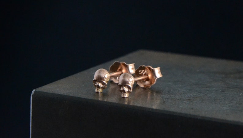 May include: A pair of rose gold skull stud earrings. The earrings are small and delicate, and they feature a detailed skull design.