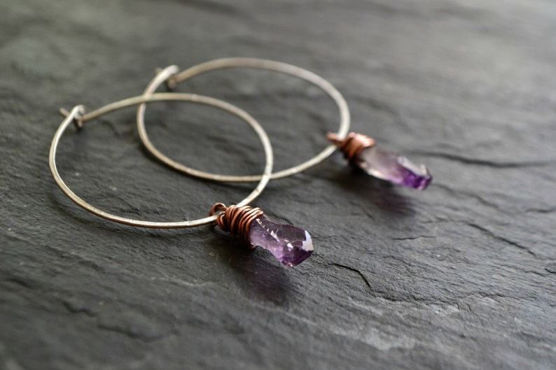 May include: A pair of silver hoop earrings with amethyst crystal accents wrapped in copper wire.