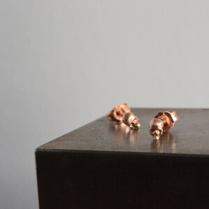 May include: Three rose gold skull stud earrings on a dark gray surface.