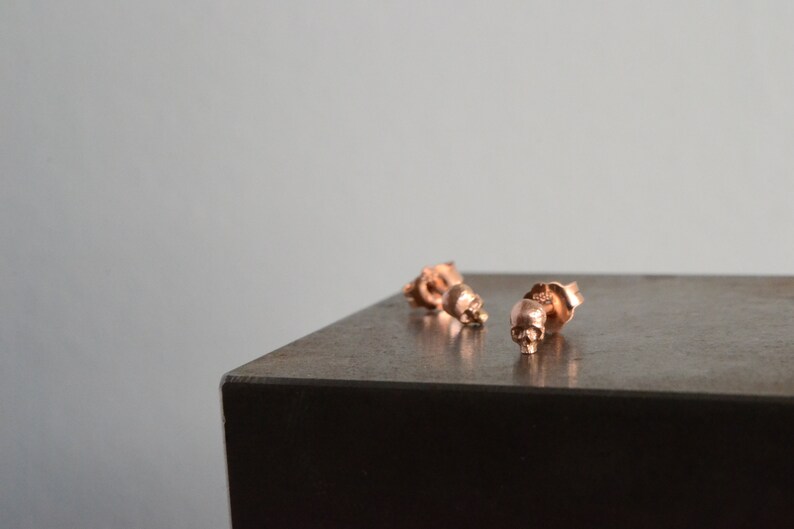 May include: Three rose gold skull stud earrings on a dark gray surface.