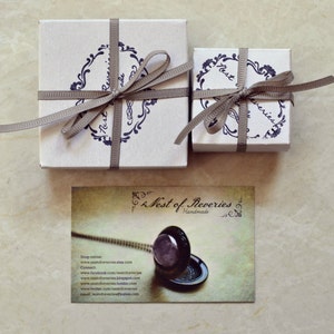 May include: Two small gift boxes wrapped in white paper with brown ribbon and a blue and white floral design. The boxes are tied with a bow. The smaller box has the text "Nest of Reveries" on it. The larger box has the text "Nest of Reveries Handmade" on it. A business card with the text "Nest of Reveries Handmade" is also shown.