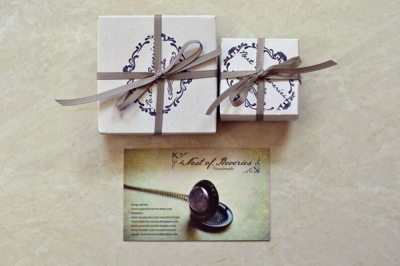 May include: Two small gift boxes wrapped in white paper with brown ribbon and a blue and white floral design. The boxes are tied with a bow. The smaller box has the text "Nest of Reveries" on it. The larger box has the text "Nest of Reveries Handmade" on it. A business card with the text "Nest of Reveries Handmade" is also shown.