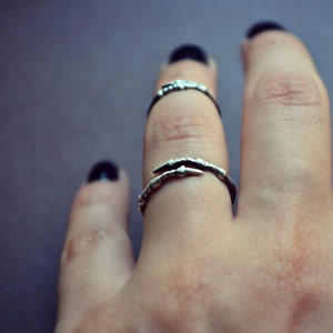 Könnte beinhalten: Ein silberner Ring mit einem Krallen-Design. Der Ring wird am Mittelfinger einer Hand mit schwarzem Nagellack getragen.