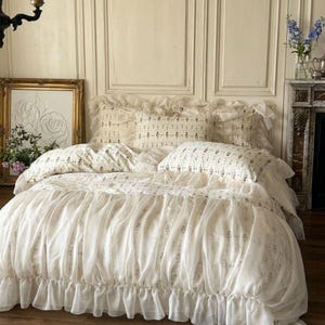 May include: A cream-colored bed with a ruffled duvet cover and matching pillow shams. The bedding has a delicate floral pattern and is set against a paneled wall. A framed artwork and flowers in a vase are also visible.