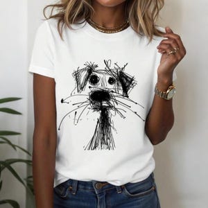 May include: White t-shirt featuring a whimsical black and white sketch of a dog's face. The dog's features are created with expressive, scribbled lines. The shirt is a classic crew neck style.