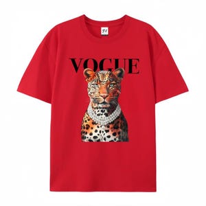 May include: Red t-shirt featuring a leopard wearing a diamond necklace. The word "VOGUE" is printed in black above the leopard's head. The shirt has short sleeves and a crew neck.