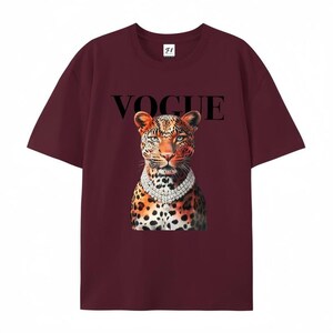 May include: Burgundy t-shirt featuring a leopard illustration wearing a diamond necklace, with the word "VOGUE" above the leopard's head. The shirt has short sleeves and a crew neck.