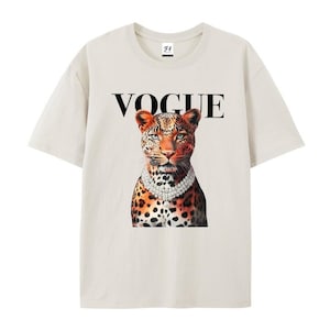 May include: Cream-colored t-shirt featuring a leopard illustration wearing a diamond necklace, with the word "VOGUE" in black above the leopard. The leopard has orange and black spots and blue eyes. The shirt has short sleeves and a crew neck.