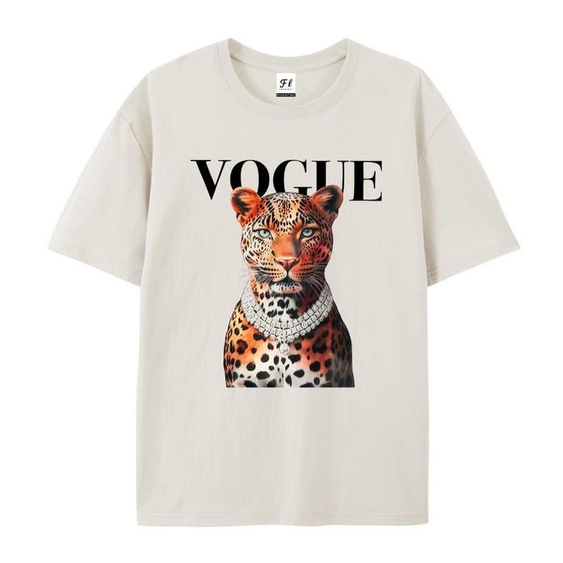May include: Cream-colored t-shirt featuring a leopard illustration wearing a diamond necklace, with the word "VOGUE" in black above the leopard. The leopard has orange and black spots and blue eyes. The shirt has short sleeves and a crew neck.
