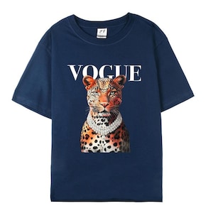 May include: Navy blue t-shirt featuring the word "VOGUE" in white above a leopard wearing a diamond necklace. The leopard has orange and black spots and is centered on the shirt.