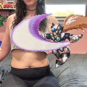 Könnte beinhalten: Pixel-Art-Kreation eines Charakters, der eine halbmondförmige Waffe hält. Die Waffe weist Schattierungen von Lila und Weiß auf, wobei der Charakter in Schwarz, Weiß und Orange gehalten ist. Das Kunstwerk besteht aus kleinen, farbigen Perlen.