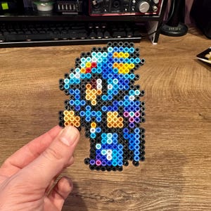 May include: A pixel art figure made from colorful plastic beads. The figure is primarily blue, with accents of yellow, red, orange, and black. The figure is held in a person's hand against a wooden surface.