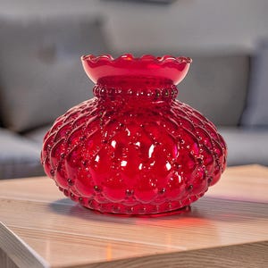 May include: A vibrant red glass vase featuring a rounded, bulbous body with a textured hobnail pattern and a ruffled, scalloped rim. The translucent glass reflects light across its surface, highlighting the intricate raised dot design.