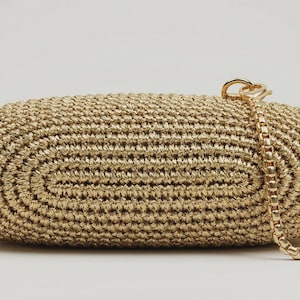 May include: An oval-shaped clutch bag crafted from metallic gold woven cord. The bag features a textured, concentric stitch pattern and includes a detachable gold-tone metal box chain strap attached to a circular ring on the top right side.