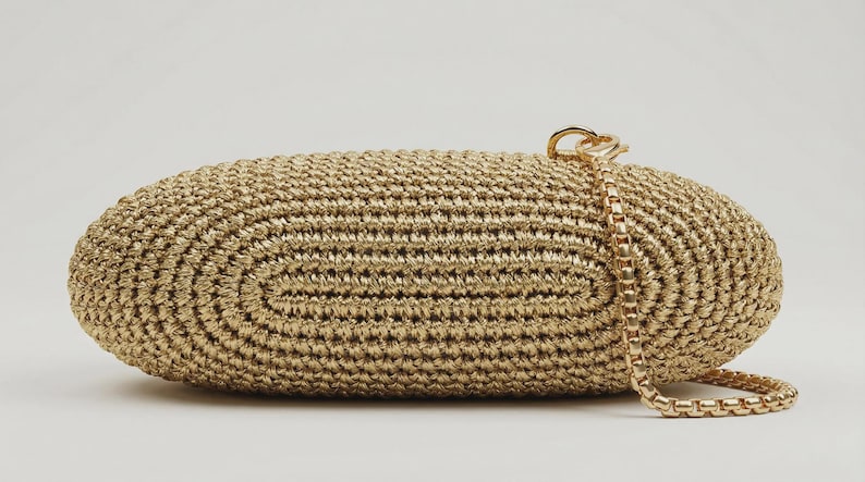 May include: An oval-shaped clutch bag crafted from metallic gold woven cord. The bag features a textured, concentric stitch pattern and includes a detachable gold-tone metal box chain strap attached to a circular ring on the top right side.