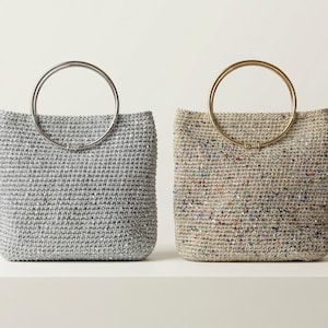 Puede incluir: Dos bolsos cuadrados tejidos con asas circulares. Uno es gris con un asa plateada y el otro beige con un asa dorada. Los bolsos se exhiben sobre una superficie blanca sobre un fondo blanco.