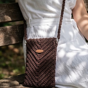 May include: A brown macrame crossbody phone bag with a chevron pattern. The bag has a long, braided strap and a small, rectangular tag that reads "Hand Made". The bag is hanging from a silver ring.