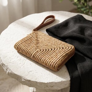 May include: A woven straw clutch with a brown leather wrist strap. The rectangular bag features a textured, wave-like pattern and a brown zipper. A black fabric is draped nearby on a white stone surface.