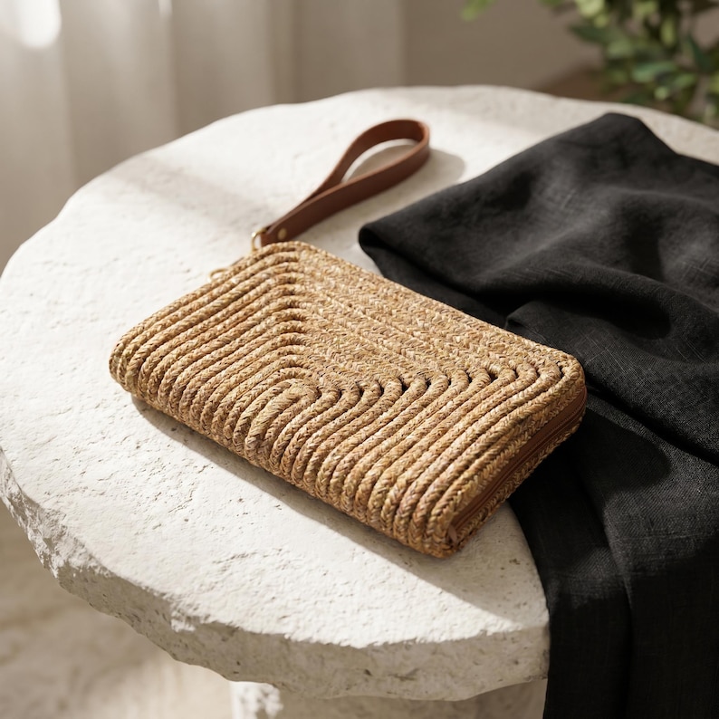 May include: A woven straw clutch with a brown leather wrist strap. The rectangular bag features a textured, wave-like pattern and a brown zipper. A black fabric is draped nearby on a white stone surface.