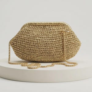 May include: A gold-toned crochet clutch bag with a structured frame closure and a matching gold-toned metal chain shoulder strap. The bag features a textured, woven surface with a metallic sheen, designed in a rounded, elongated shape.