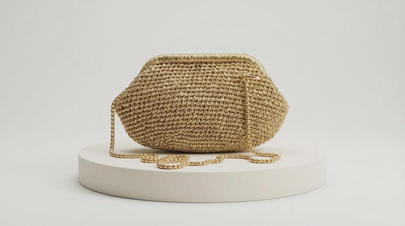 May include: A gold-toned crochet clutch bag with a structured frame closure and a matching gold-toned metal chain shoulder strap. The bag features a textured, woven surface with a metallic sheen, designed in a rounded, elongated shape.