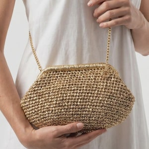May include: A gold-colored woven clutch bag with a textured, straw-like appearance. The bag features a metallic gold frame with a hinged closure and a thin, gold-toned metal chain strap attached to the sides for carrying over the shoulder.