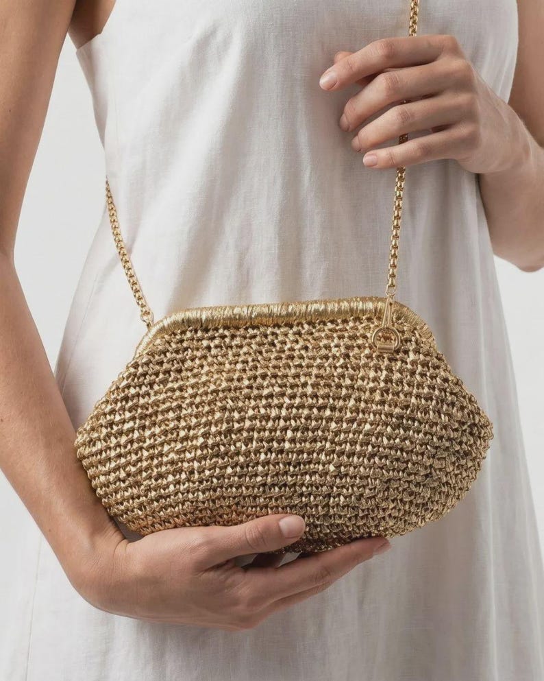 May include: A gold-colored woven clutch bag with a textured, straw-like appearance. The bag features a metallic gold frame with a hinged closure and a thin, gold-toned metal chain strap attached to the sides for carrying over the shoulder.