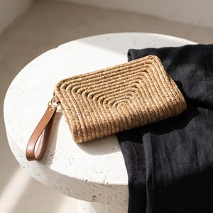 May include: A woven straw clutch with a brown leather wrist strap. The rectangular bag features a geometric design and a gold-colored clasp. The clutch is tan and brown, and rests on a white surface next to a black fabric.