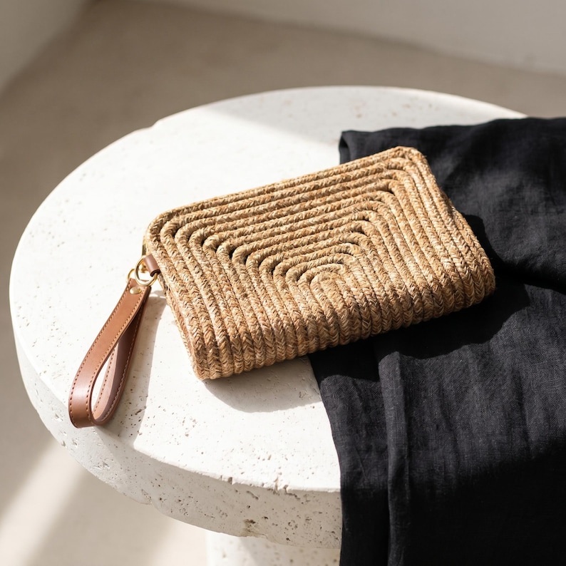 May include: A woven straw clutch with a brown leather wrist strap. The rectangular bag features a geometric design and a gold-colored clasp. The clutch is tan and brown, and rests on a white surface next to a black fabric.