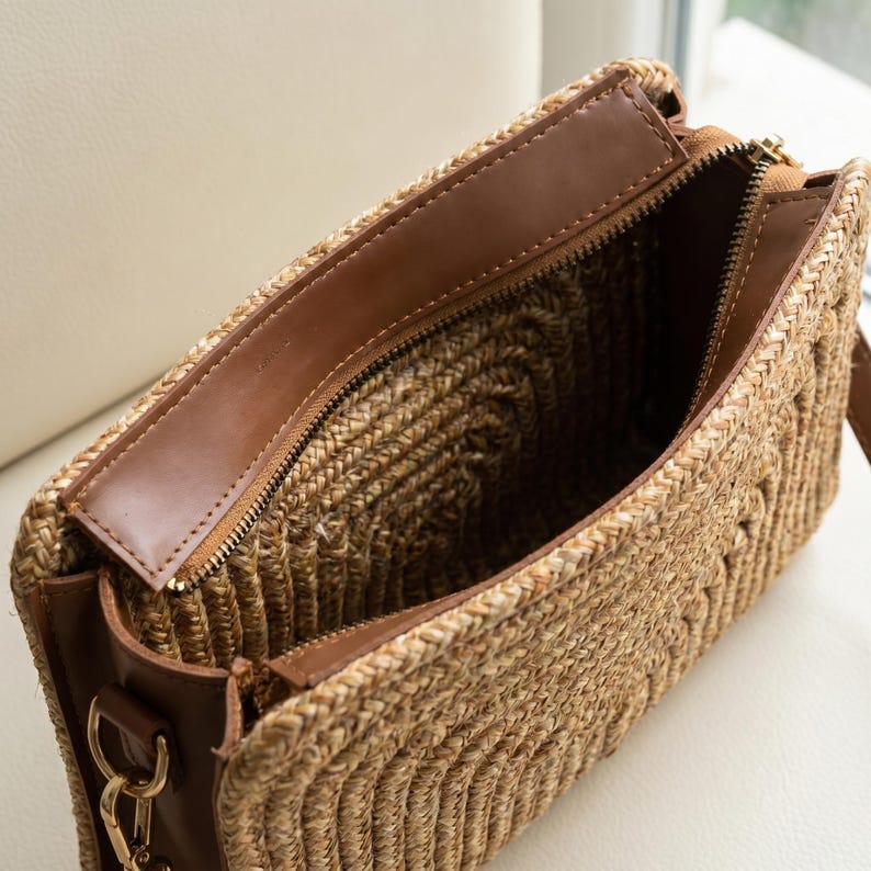 May include: Open view of a woven straw handbag with a brown leather trim and zipper. The interior is lined with a dark fabric. The bag has a long, adjustable strap and gold-colored hardware.