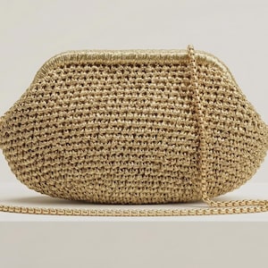 May include: A gold-colored woven clutch bag with a structured frame closure and a matching gold-toned chain shoulder strap. The bag features a textured, crocheted-style surface with a metallic sheen, shaped in a rounded, wide silhouette.