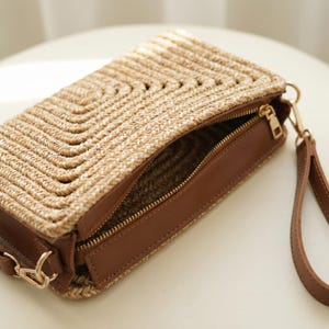 May include: A rectangular straw handbag with a woven chevron pattern on the top flap. The bag features a brown leather base and trim, a gold zipper, and a matching wrist strap. The interior is visible through the open zipper.